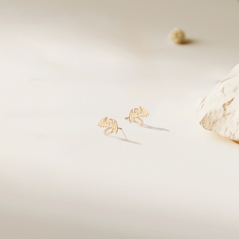 May include: A pair of gold dragon stud earrings. The earrings are small and delicate, and the dragons are facing forward with their wings spread out.