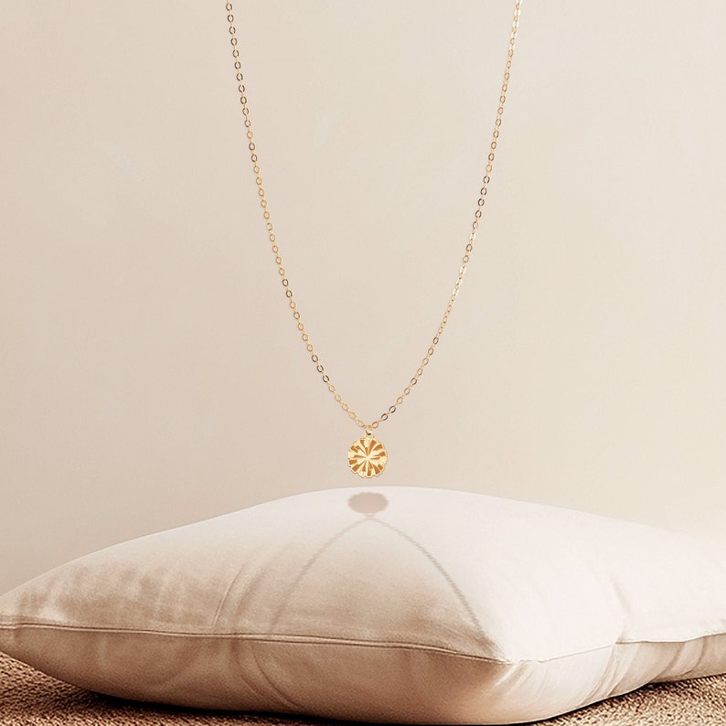 May include: A delicate gold necklace with a circular pendant featuring a sunburst design. The pendant hangs from a fine chain, creating a minimalist and elegant look. The necklace is displayed against a neutral background.