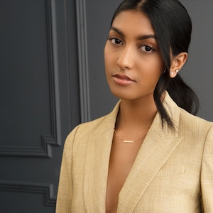 May include: A woman with dark hair wearing a light brown blazer with a gold necklace. She is looking directly at the camera.