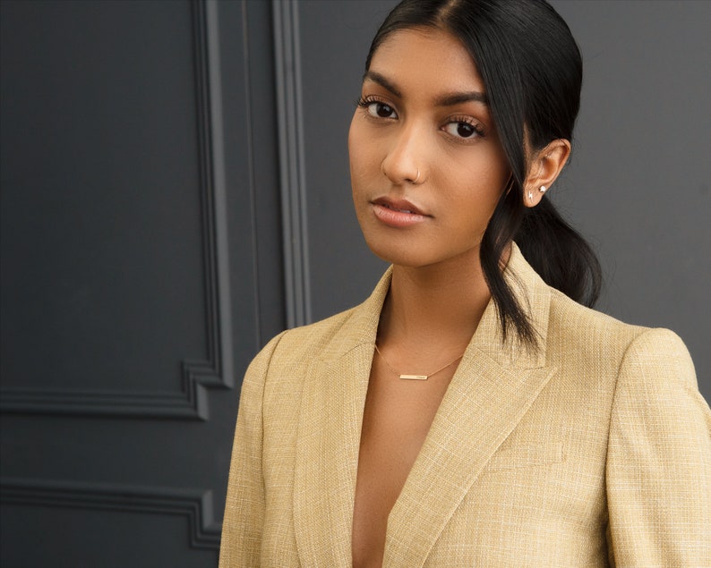 May include: A woman with dark hair wearing a light brown blazer with a gold necklace. She is looking directly at the camera.