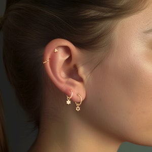 May include: A person wearing gold hoop earrings with a small flower charm and a Star of David charm.