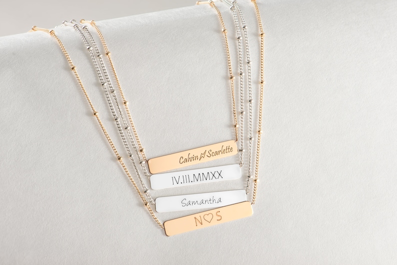May include: Four gold bar necklaces with different engravings. The top bar reads "Calvin & Scarlette", the second bar reads "IV.III.MMXX", the third bar reads "Samantha", and the bottom bar reads "NOS" with a heart symbol.