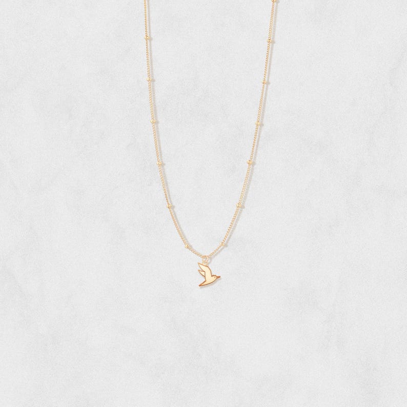 May include: A delicate gold-colored necklace featuring a small bird pendant. The necklace has a fine chain with small, evenly spaced beads. The bird pendant is a simple silhouette, adding a touch of elegance to the jewelry.