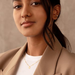 May include: A gold necklace with a small, circular pendant and a beaded chain. The necklace is worn by a person in a tan blazer and white top. The person has dark hair and a nose ring.