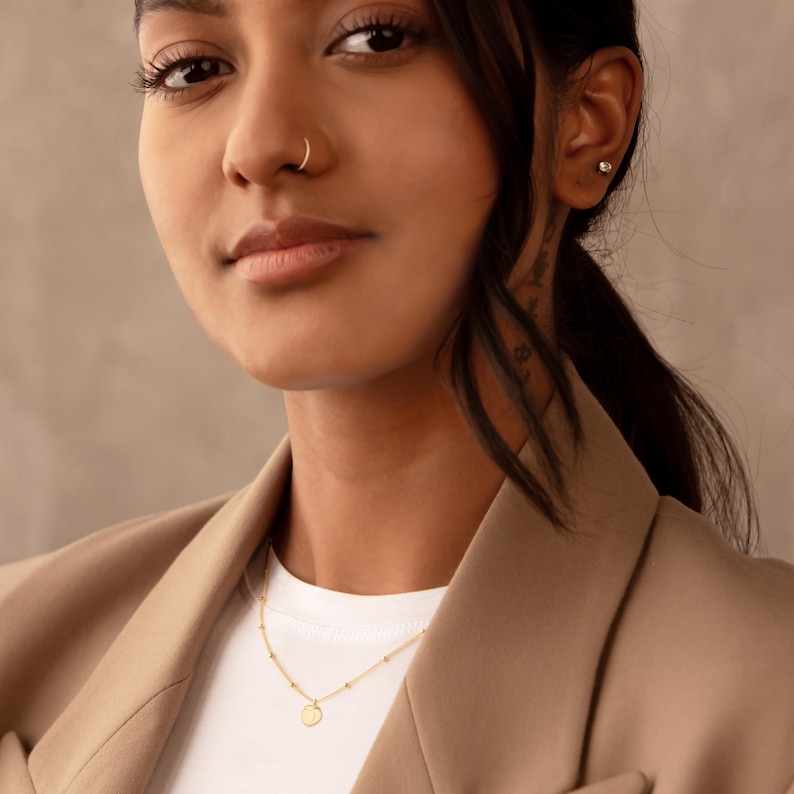 May include: A gold necklace with a small, circular pendant and a beaded chain. The necklace is worn by a person in a tan blazer and white top. The person has dark hair and a nose ring.