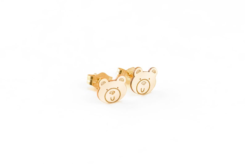 May include: A pair of gold bear-shaped stud earrings. The bears have a simple, cartoon-like design with a small nose and mouth.