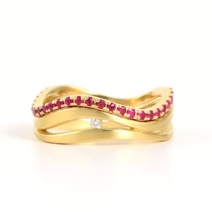 Puede incluir: Un anillo de oro con tres bandas, la banda superior está engastada con una fila de rubíes, la banda del medio tiene un solo diamante y la banda inferior es lisa.