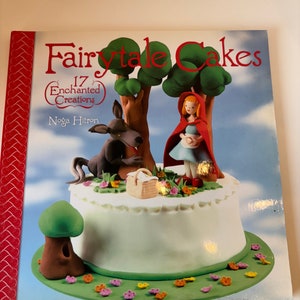 May include: A book titled "Fairytale Cakes: 17 Enchanted Creations" by Noga Hitron. The cover features a cake decorated with a red-hooded girl, a wolf, and a basket. The cake is surrounded by green grass and pink flowers.