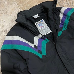 Może przedstawiać: Czarna kurtka puchowa z geometrycznym wzorem w kolorze białym, fioletowym i zielonym. Kurtka zapinana jest na zamek błyskawiczny i ma wysoki kołnierz. Etykieta na kurtce głosi "aditex" i "adidas".