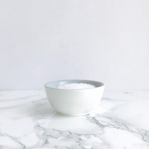 May include: A white ceramic salt cellar with a light blue rim, filled with white salt, sitting on a white marble surface.
