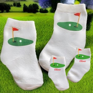 May include: White golf socks with a green golf course design, a red flag, and a golf ball. Two pairs of socks are shown, one pair with the name "Oliver" printed on them.