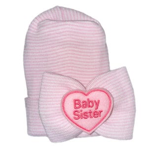Puede incluir: Un gorro de bebé a rayas rosas y blancas con un lazo grande y una aplicación en forma de corazón. La aplicación de corazón es rosa con las palabras "Baby Sister" bordadas en rosa oscuro. El gorro está diseñado para recién nacidos.