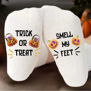 May include: White Halloween socks with the phrases "Trick or Treat" and "Smell My Feet" printed in black. The socks feature orange jack-o'-lanterns and candy baskets, perfect for the spooky season.