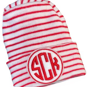 Op de afbeelding: Een rood-wit gestreepte beanie-muts met een cirkelvormige patch met de geborduurde initialen "SCB" in rood. De muts is gemaakt van een zacht, gebreid materiaal.