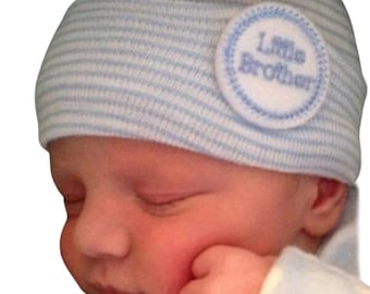 May include: A baby wearing a blue and white striped knit hat with a blue circle patch that says "Little Brother" in white embroidery.