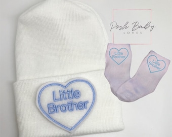May include: White baby hat with a blue heart-shaped patch that says "Little Brother".  The hat is paired with a pair of white socks with blue hearts that say "Little Brother".