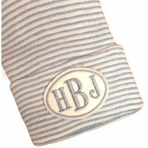 May include: A white and blue striped baby hat with a white oval patch embroidered with the initials "HBJ" in silver thread.