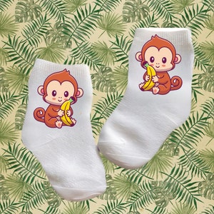 May include: A pair of white ankle socks with a cartoon monkey holding a banana on each sock. The monkey is brown with a yellow banana and has a cute expression.
