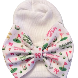 May include: A white knit beanie featuring a large fabric bow attached to the front. The bow has a white background with a repeating pattern of golf clubs, golf balls, pink flags, small pink bows, hearts, and the text Daddy's Little Golf Buddy.