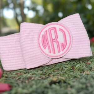 May include: A pink and white striped bow with a circular monogram patch. The monogram features the letters "MRJ" in a matching pink thread. The bow is resting on a bed of green moss.