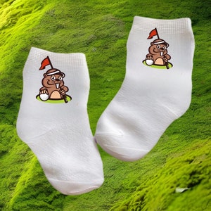 May include: A pair of white ankle socks with a cartoon image of a gopher playing golf. The gopher is wearing a hat and holding a golf club. The socks have a red, white, and green color scheme.