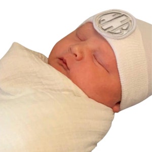 May include: A newborn baby swaddled in a cream-colored blanket, wearing a white beanie with a silver emblem. The baby is sleeping peacefully, with closed eyes and a serene expression.