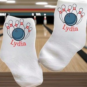 May include: White ankle socks with a bowling theme. Each sock features a blue bowling ball and red and white pins graphic, with the name "Lydia" in red script. The socks are displayed against a blurred bowling alley background.