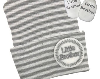 May include: Gray and white striped baby hat with a white circle patch that says "Little Brother" and two matching gray and white striped mittens with the same text.