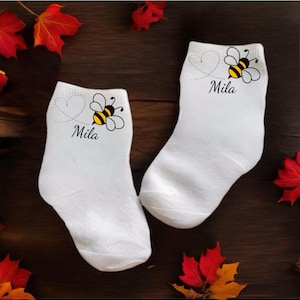 Può includere: Un paio di calzini bianchi alla caviglia con un design a forma di ape e cuore. I calzini hanno il nome "Mila" stampato su di essi.