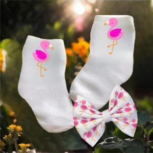 May include: A pair of white socks with pink flamingo designs and a white bow with pink flamingo designs.