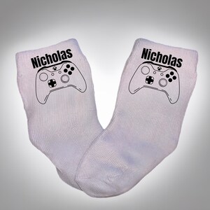 May include: A pair of white socks with a black graphic of a video game controller and the name "Nicholas" printed above the controller. The socks are made of a ribbed knit fabric.