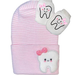 Könnte beinhalten: Rosa-weiß gestreiftes Baby-Mützchen mit einem weißen Zahnapplikation mit einer rosa Schleife. Die Mütze wird mit einem Paar weißen Fäustlingen mit Zahnapplikationen und rosa Schleifen geliefert.