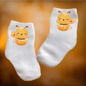 Può includere: Un paio di calzini bianchi alla caviglia con un simpatico design di ape di cartone animato. L'ape tiene un favo di miele ed è seduta in un barattolo di miele.