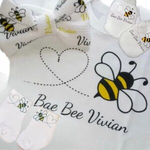 May include: A white baby onesie with a bee and heart design. The onesie has the text "Bae Bee Vivian" on it. The set also includes a matching bow tie and a pair of booties.