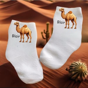 May include: Pair of white ankle socks featuring a cartoon camel illustration with the name "Blair" printed in black. The camel is tan with a brown hump. The socks are set against a desert backdrop.