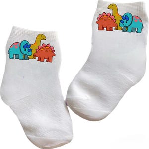 May include: A pair of white socks with a dinosaur graphic. The socks feature a blue, red, and yellow dinosaur with a green dinosaur in the background.