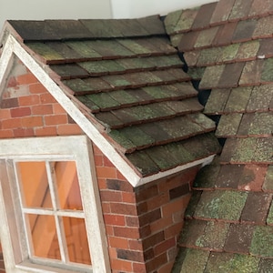 Op de afbeelding: Een miniatuur bakstenen gebouw met een groen en bruin pannendak. Het dak heeft een verweerde uitstraling. Een wit omrand raam is zichtbaar aan de zijkant van het gebouw. Het metselwerk is rood en het raam heeft een oranje gloed.