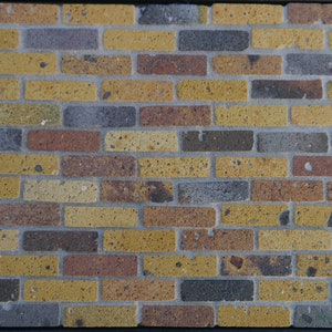 May include: A close-up of a brick wall with a variety of colors. The bricks are primarily yellow, with accents of gray, brown, and red. The mortar between the bricks is a light gray color. The wall has a rough, textured surface.