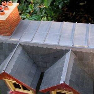 May include: A model of a house with a grey slate roof and a chimney. The roof is made of individual slate tiles and has a decorative edge.