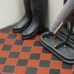 May include: A black metal umbrella stand with a gray umbrella and a pair of black rain boots. The stand has a rectangular base with a curved top and two compartments. The stand is sitting on a black and red checkered floor.