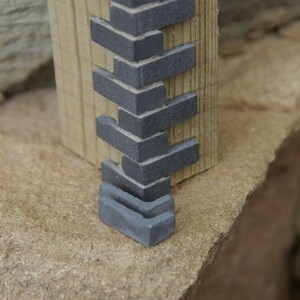 May include: A close-up of a miniature brick wall made of gray bricks. The bricks are stacked in a staggered pattern and are attached to a wooden beam.
