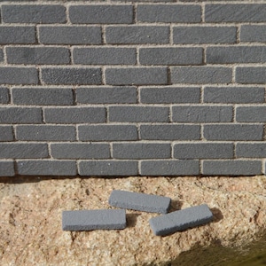 May include: Three gray brick-shaped blocks, possibly for model building or crafts, are sitting on a rough, brown surface. The blocks are rectangular and have a slightly textured surface.