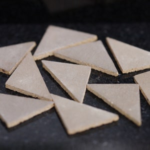 May include: A collection of twelve triangular ceramic tiles in a light beige colour. The tiles are arranged in a random pattern on a black surface.