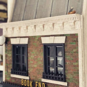 May include: A miniature brick building with two windows and a sign that reads "BOOK END". The building has a white cornice with decorative details and a brown roof.