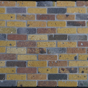 May include: A brick wall with a mix of yellow, brown, and gray bricks. The bricks have a rough texture and are arranged in a staggered pattern.