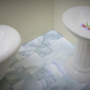 Puede incluir: Un lavabo de pedestal de porcelana blanca con un diseño floral y detalles dorados se encuentra sobre un suelo de baldosas. Una bañera blanca está en el fondo.