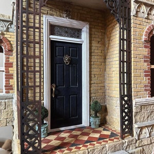May include: A black front door with a lion's head knocker and a decorative glass window above. The door is framed by a white trim and is set within a brick wall with a trellis overhead. Two small potted plants flank the door.