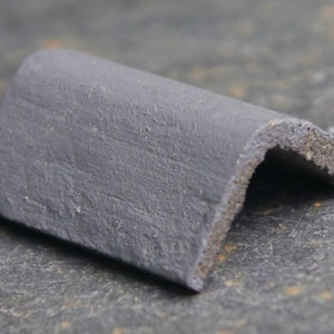 Puede incluir: Una teja de esquina de color gris oscuro para un tejado. La teja está hecha de un material texturizado y tiene una forma curva.