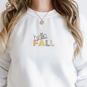 Puede incluir: Sudadera blanca con el texto "hello fall" en rayas marrones, amarillas y naranjas.
