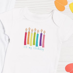 May include: White baby onesie with a rainbow of candles and the text "It's my birthday".